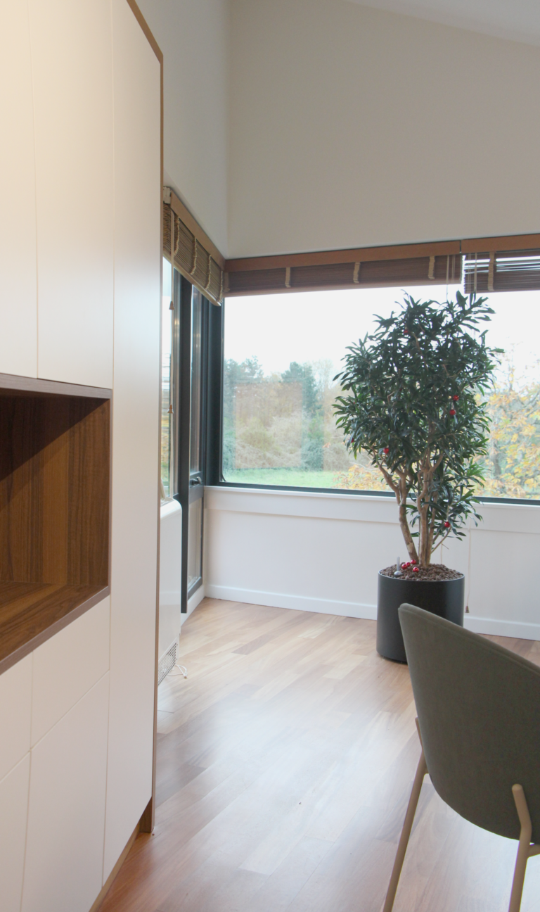 Coin bureau lumineux avec une grande fenêtre, une plante en pot et un meuble de rangement en bois et blanc.