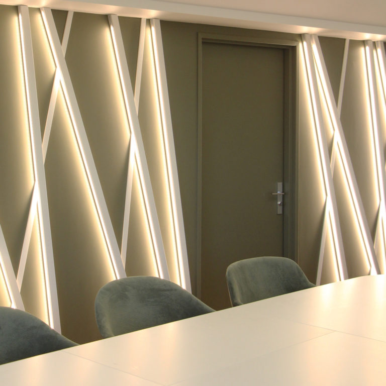 Mur décoratif avec des bandes lumineuses en motifs diagonaux et des chaises en velours vert autour d'une table blanche.