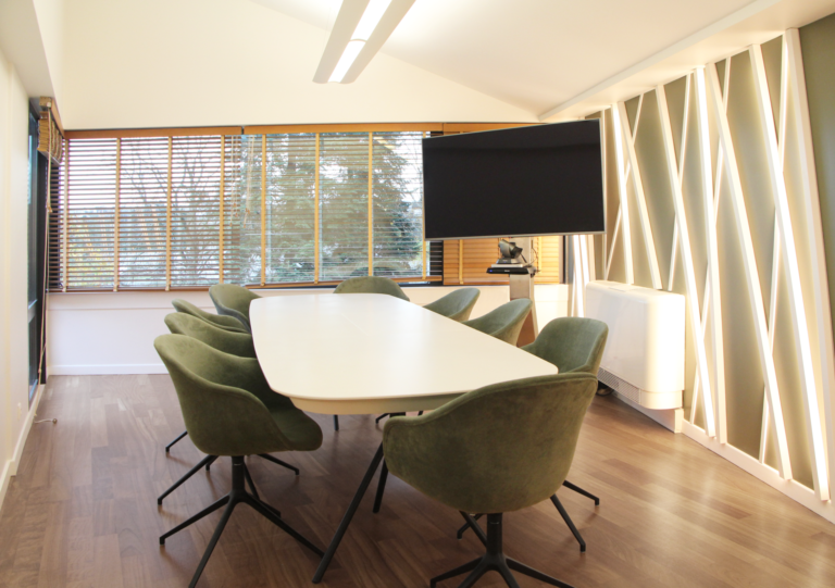 Salle de réunion lumineuse avec une grande table blanche, des chaises vertes en velours, un mur décoratif en bois et des stores en bois aux fenêtres.