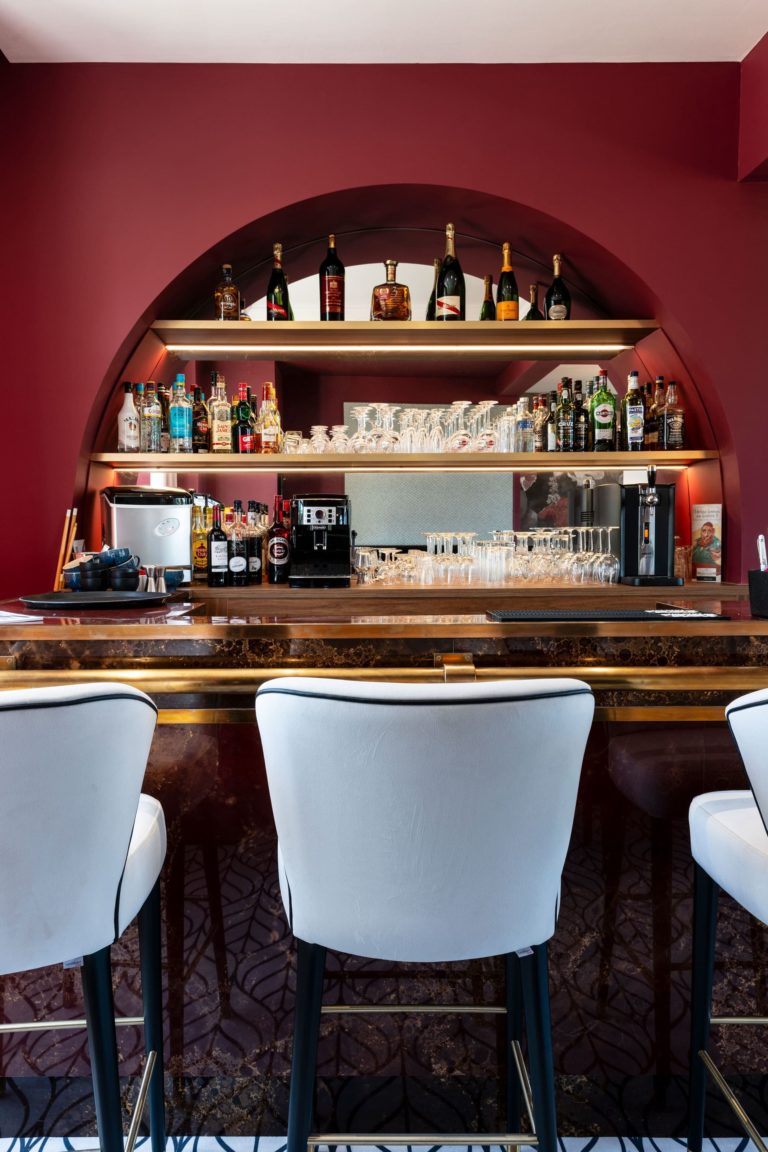 Bar rénové d'un hôtel des années 30 avec des murs bordeaux et des accents dorés, conçu par JoyDesign Intérieur.