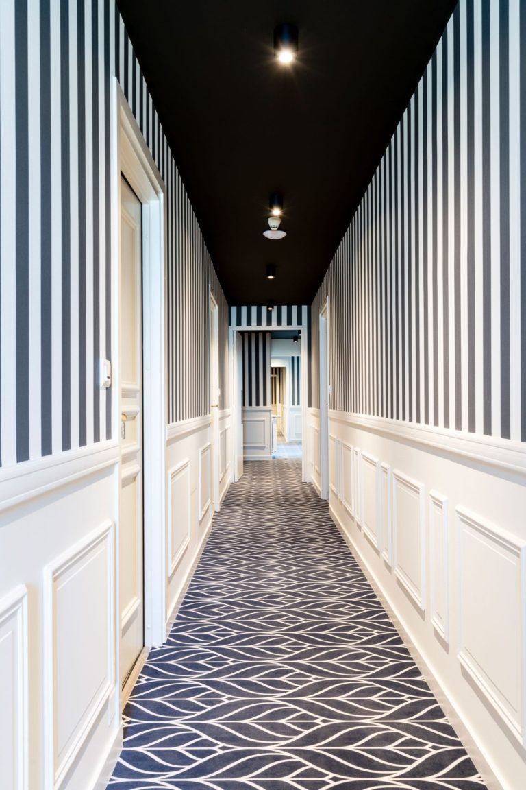 Couloir rénové d'un hôtel des années 30 avec des rayures verticales noir et blanc et des moulures blanches, conçu par JoyDesign Intérieur.