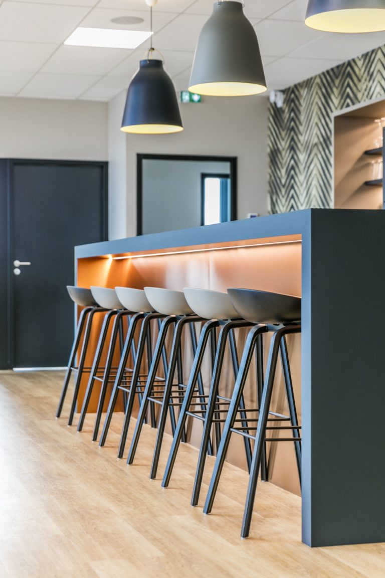 Détail d'un bar moderne avec des tabourets hauts et des luminaires suspendus dans des bureaux, conçu par JoyDesign Intérieur.