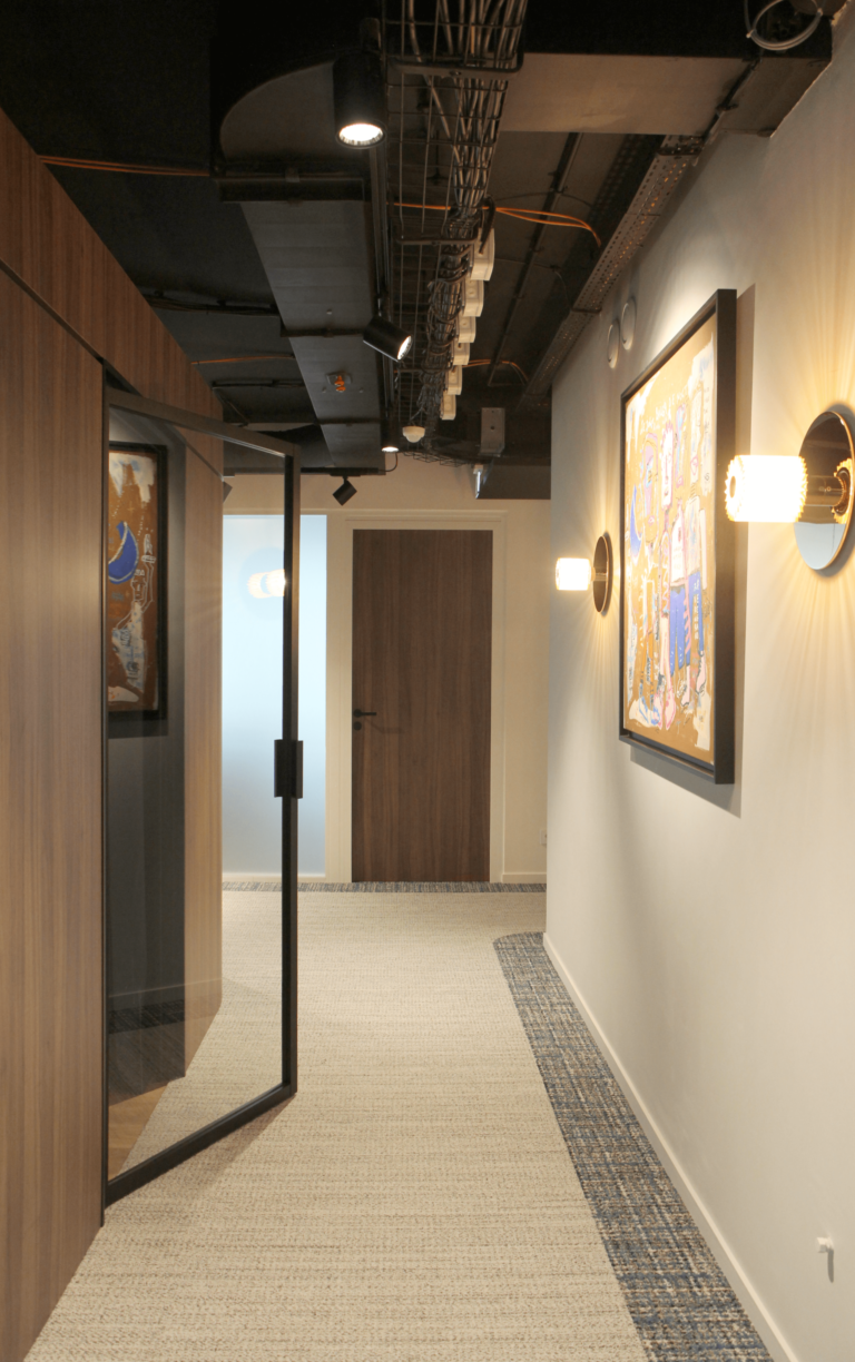 Un couloir élégant avec un plafond industriel et des murs ornés de tableaux, menant à plusieurs espaces fonctionnels dans des bureaux de 700m² conçus par JoyDesign Intérieur.