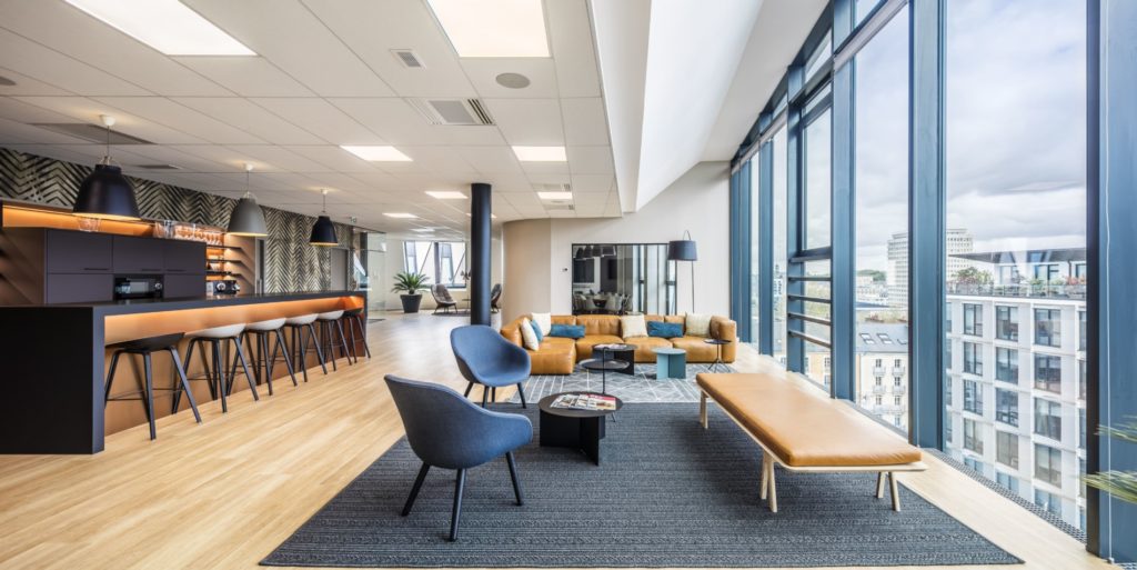 Espace de bureaux modernes avec un coin détente lumineux, un bar et une vue panoramique sur la ville, conçu par JoyDesign Intérieur.