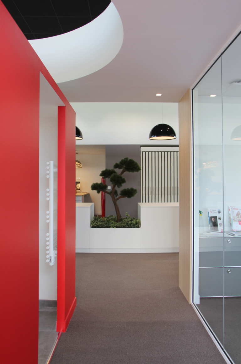 Photo d'un intérieur moderne et élégant d'une agence d'architecture, avec des murs rouges vifs, des luminaires suspendus noirs et une plante verte en pot intégré.