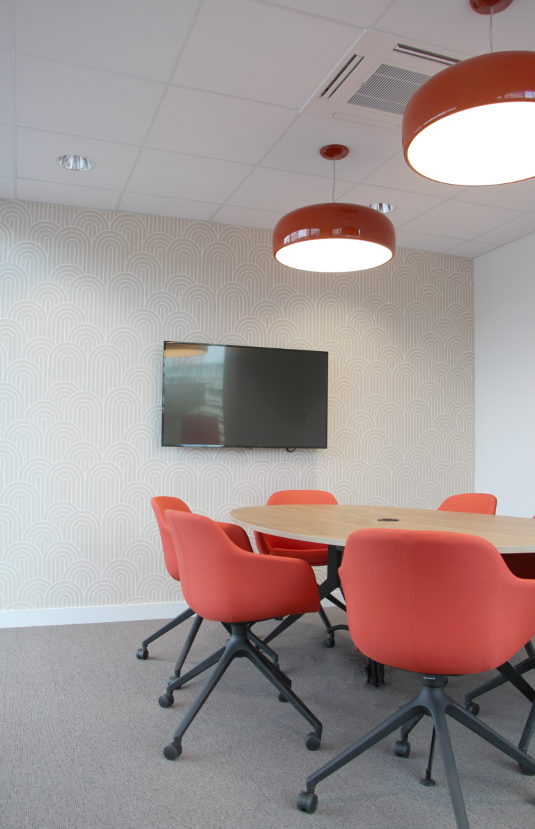 Salle de réunion moderne avec des chaises rouges, une table ovale en bois, des luminaires suspendus et un mur avec un motif géométrique.