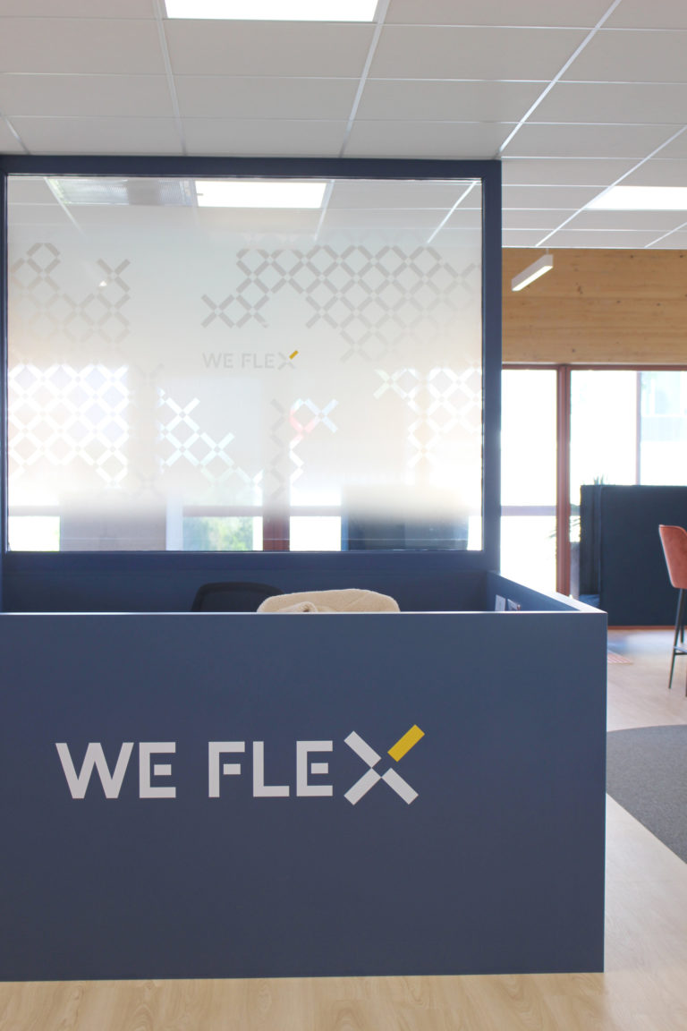 Réception moderne avec un comptoir bleu marqué "WE FLEX" et une cloison en verre partiellement givrée.