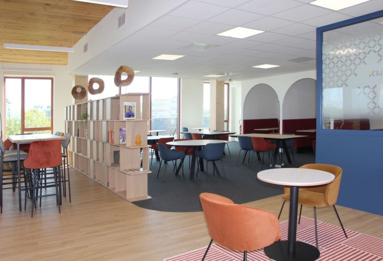 Espace de cafétéria avec des chaises colorées, des tables en bois, une étagère ouverte et des fenêtres du sol au plafond.