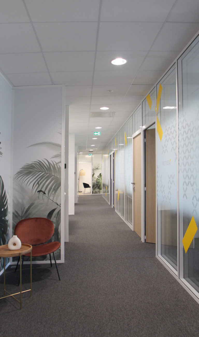 Couloir moderne de bureau avec des murs en verre, des motifs graphiques, un fauteuil orange et un papier peint à motifs végétaux.