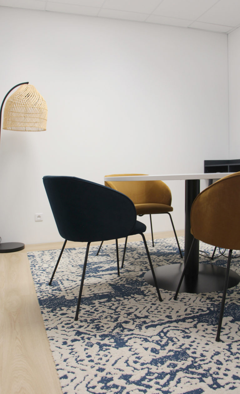 Salle de réunion moderne avec des chaises en velours bleu et jaune, une table ronde blanche et un tapis à motifs bleus et blancs.