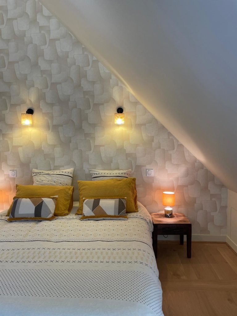 Chambre sous combles avec lit, décorée en jaune et blanc, et éclairée par des appliques murales.