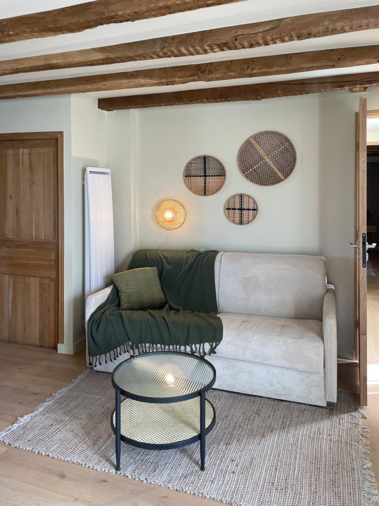 Coin salon dans une chambre avec poutres en bois apparentes, canapé beige, table basse et décoration murale en osier.