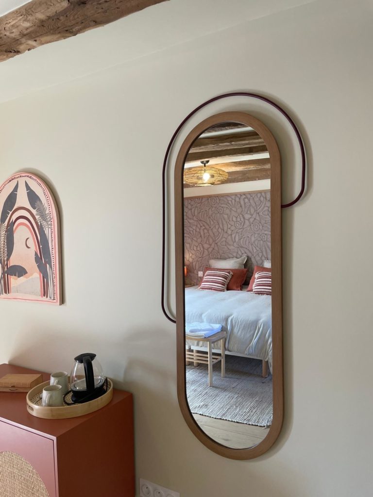 Miroir design avec cadre en bois et décoration murale, reflétant une chambre avec lit et coussins décoratifs.