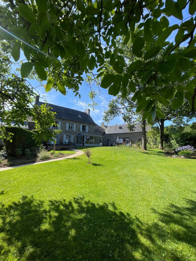 Jardin verdoyant d'une maison en pierre avec arbres et pelouse bien entretenue.