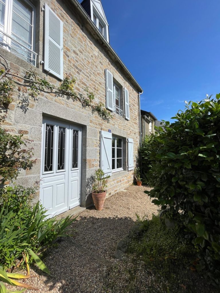 Façade d'une maison en pierre avec volets blancs et entrée avec porte vitrée, entourée de végétation.