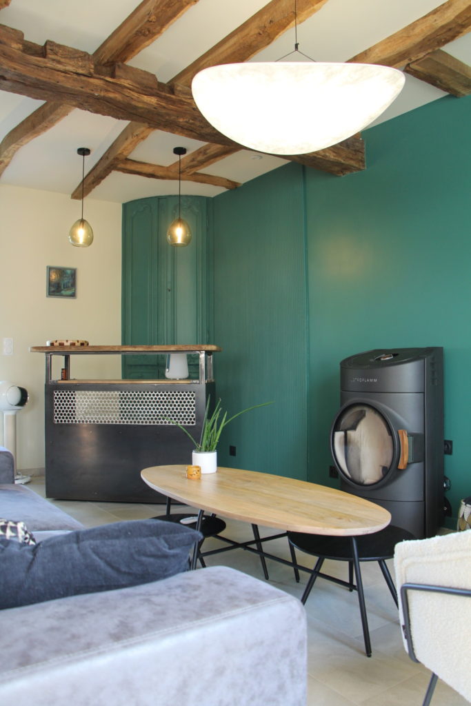 Salon moderne avec mur vert, poutres en bois apparentes, poêle à bois et bar industriel.